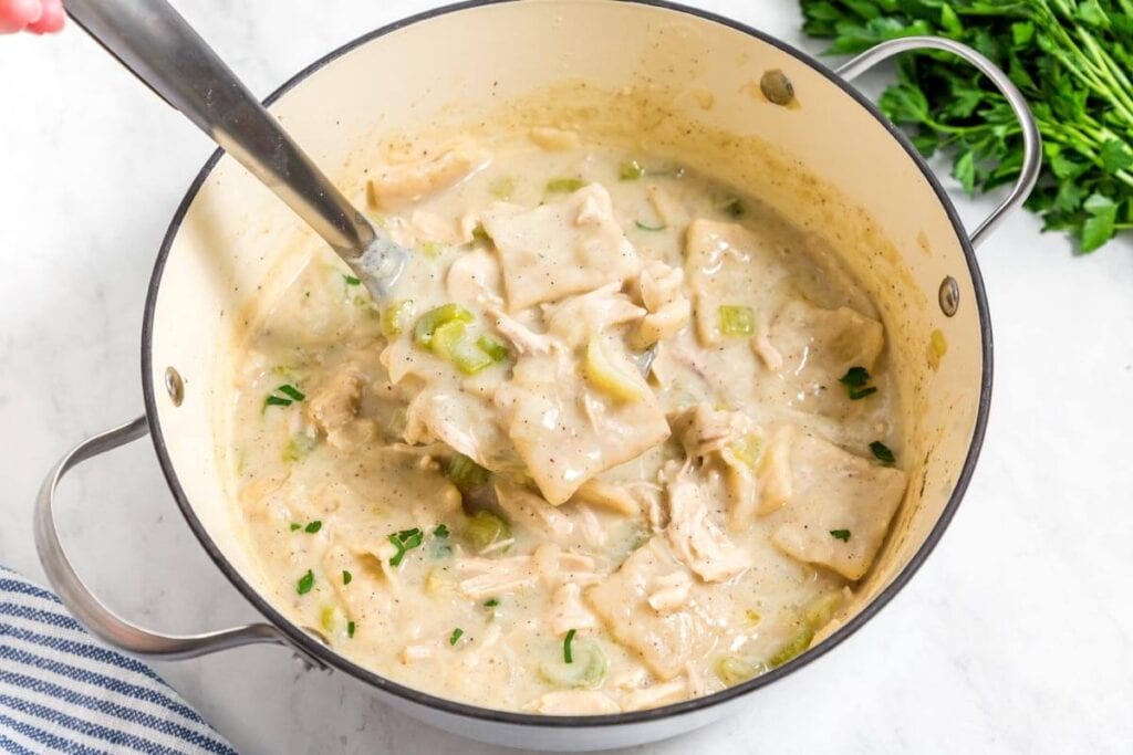 A pot of creamy chicken and dumplings with herbs and chunks of chicken, being stirred with a ladle. Fresh parsley is on the side, and a striped cloth is visible at the bottom left.