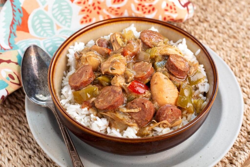 A bowl of gumbo with rice, sausages, chicken, and vegetables, served on a plate with a spoon. The dish is colorful, with peppers and herbs visible, and is placed on a woven surface with a patterned fabric in the background.